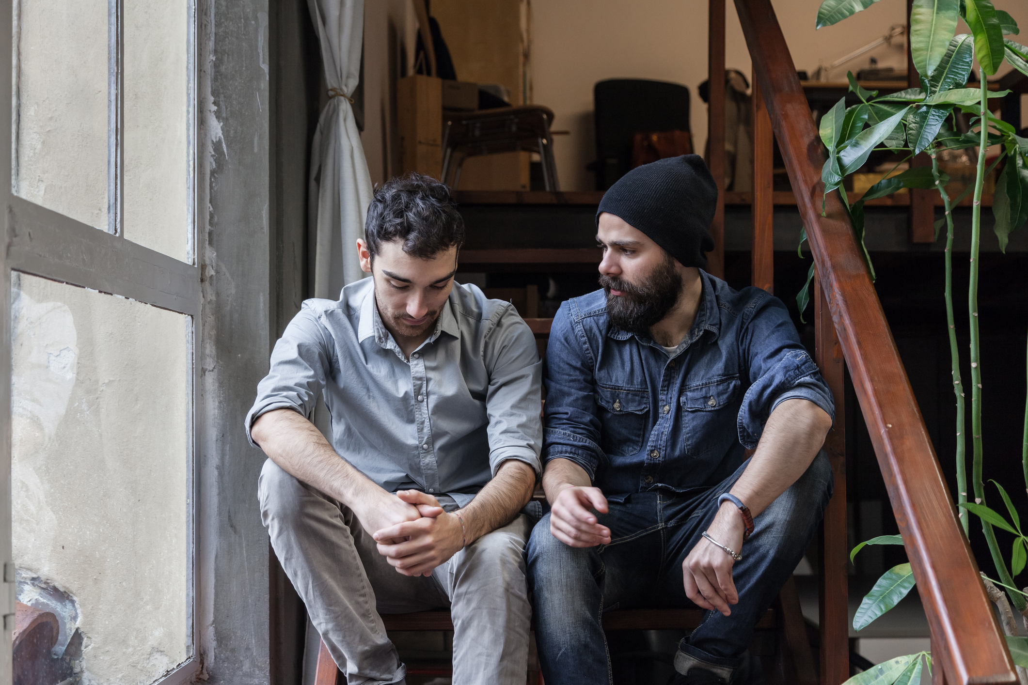 zwei junge Männer reden sitzend auf Treppe neben Grünpflannze und Fenster