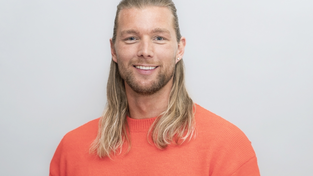 lächelnder Mann mit langen blonden Haaren und orangenem Pullover